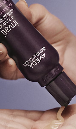 A person dispenses a cream-colored product from a purple AVEDA tube into their palm.