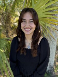 A young woman with medium-length brown hair, wearing a black dress and a pendant necklace, smiles while standing in front of green palm leaves outdoors.