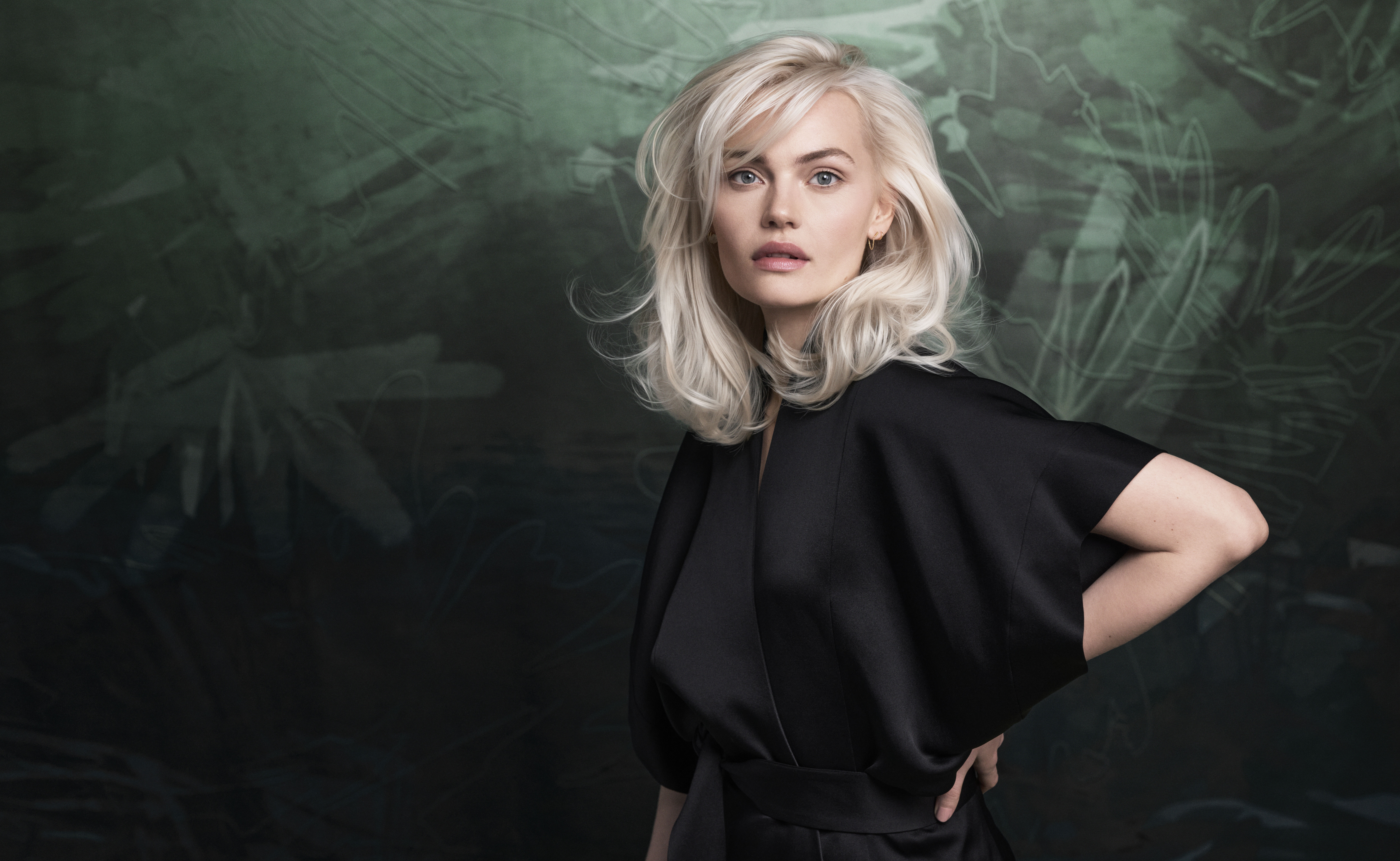 A woman with platinum blonde hair wears a black outfit, standing against a dark green abstract background with her hand on her hip. - Get Panache, FL