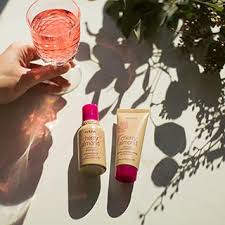 A hand holds a glass of pink liquid next to two tubes of skincare products, showing love for healthy skin, on a white surface with plant shadows.