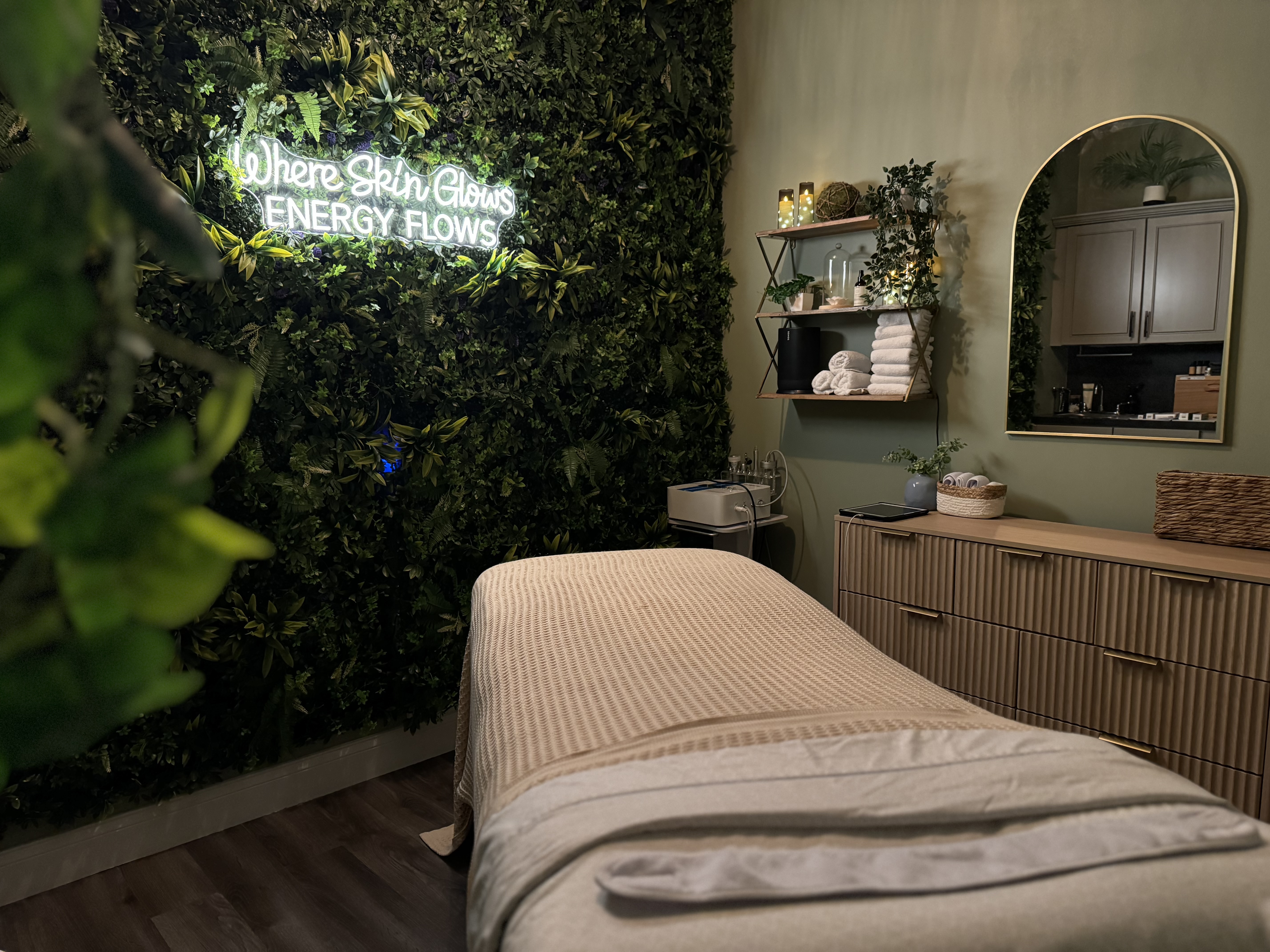 A cozy massage room at Salon Madeleine features a treatment table, a green plant wall with a neon sign reading "Where Skin Glows Energy Flows," a mirror, towels, soft lighting, and soothing Head Spa treatments.