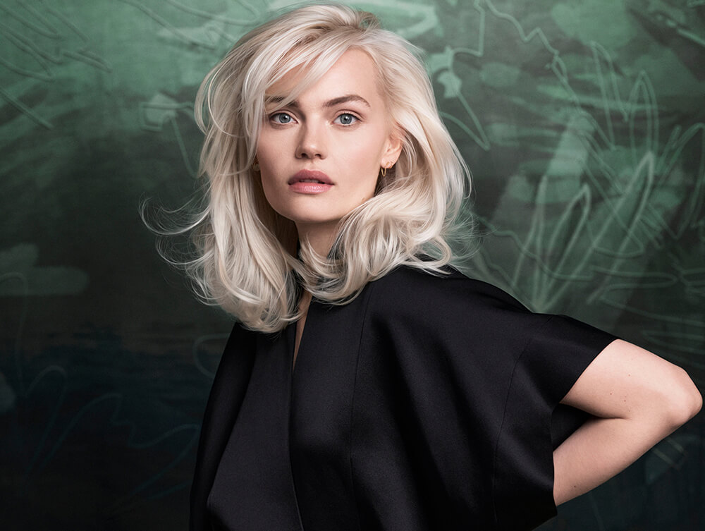 Woman with platinum blonde hair and a black top stands in front of a dark green abstract background, looking at the camera.